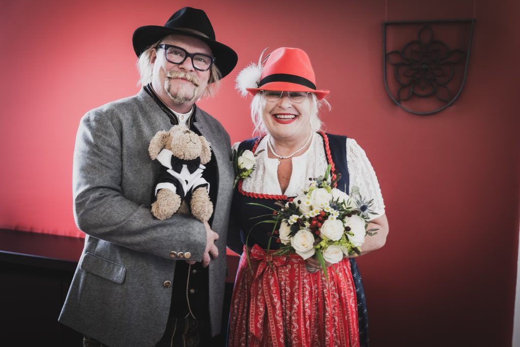 Vor dem roten Hintergrund mit Maskottchen posiert das Brautpaar - Hochzeitsfotografie Rosenheim, München und Chiemsee.